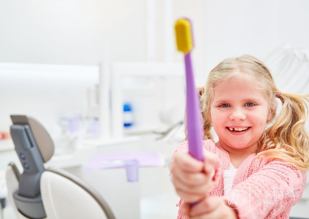 Girl in dentist's office holding up a purple toothbrush with a yellow head, smiling at the camera.