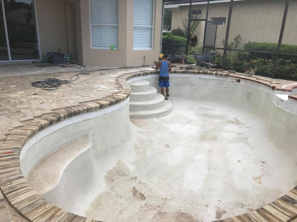 Man in pool, resurfacing it. Empty pool with stairs, brick border, beige siding and back patio.