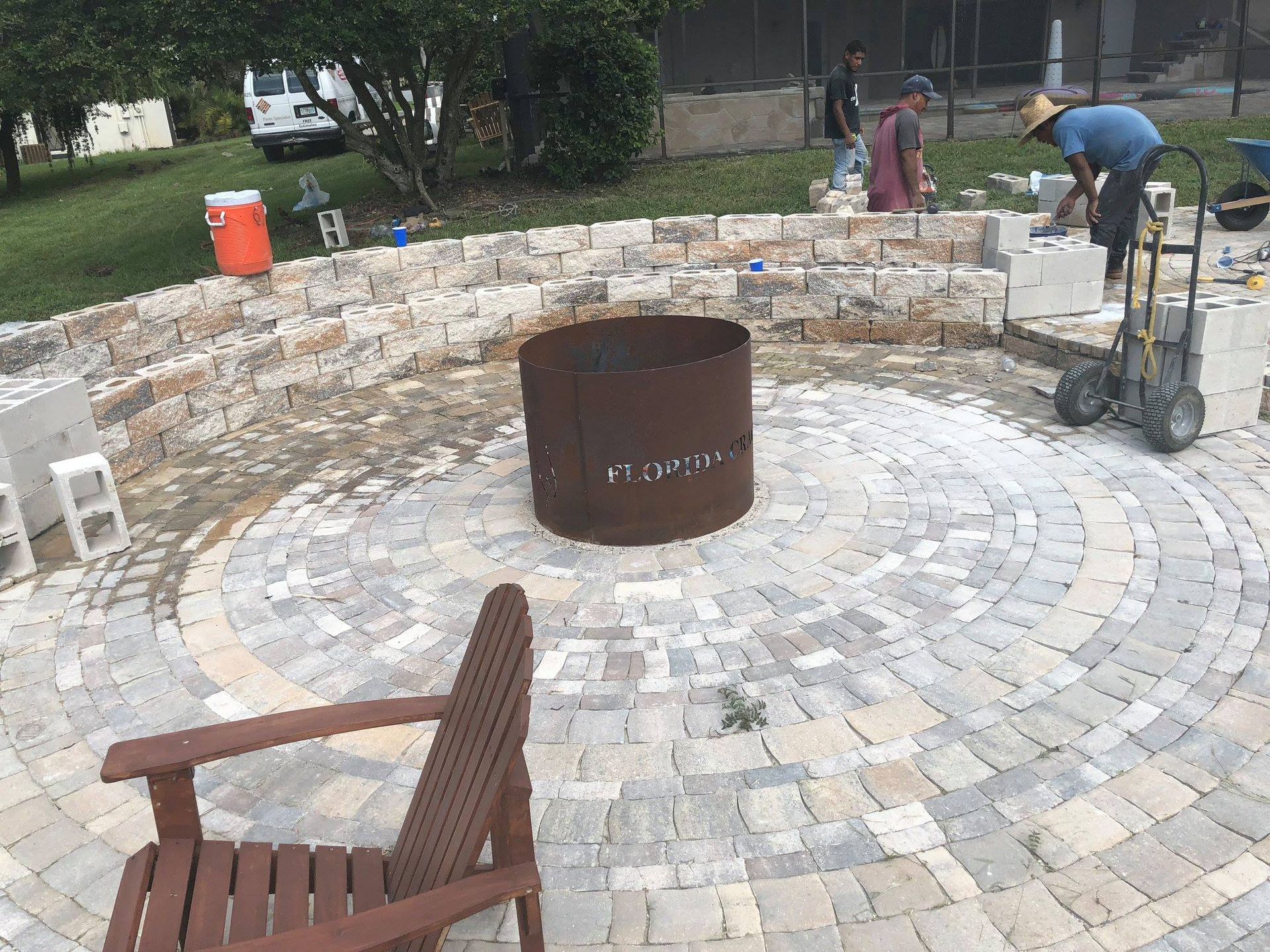 Outdoor patio under construction with a fire pit; workers laying brick pavers.