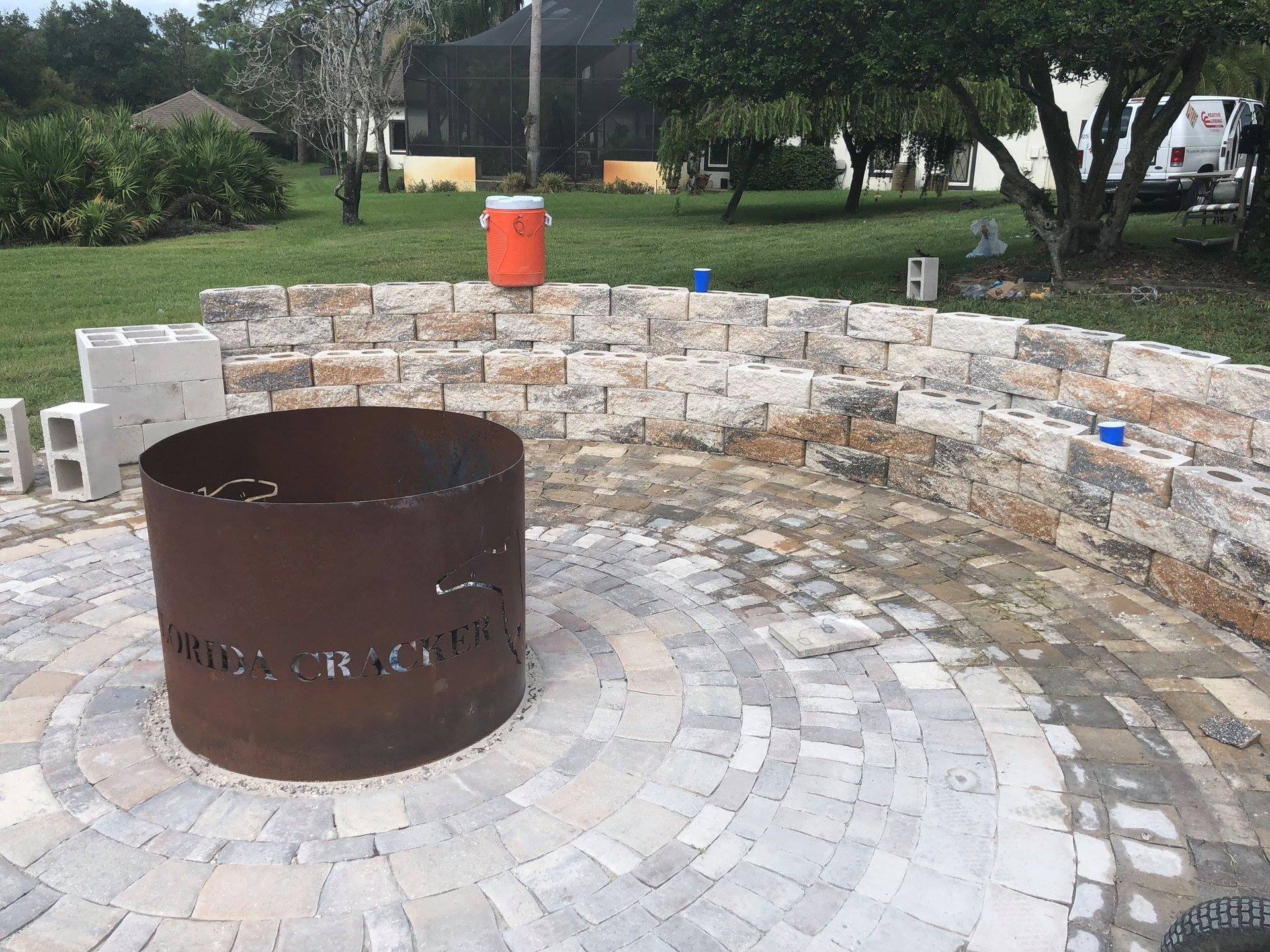 Fire pit with brick seating in a circular paved patio, with an orange cooler in the background.