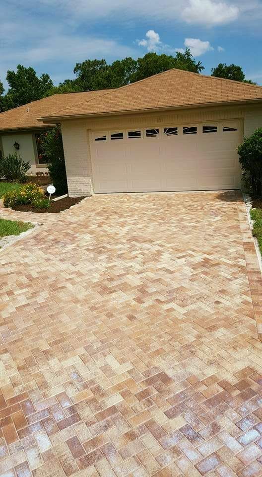 Brick driveway leads to a house with beige siding and tan roof. The garage door is closed.