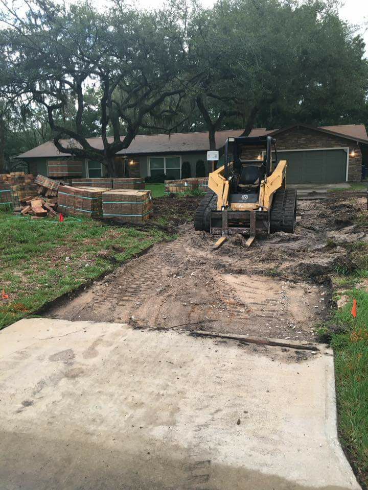 Yellow skid steer on a dirt driveway, partially demolished, in front of a house, construction in progress.