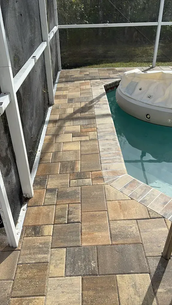 Paver patio next to a pool with a beige hot tub covered. A screen enclosure borders the patio.