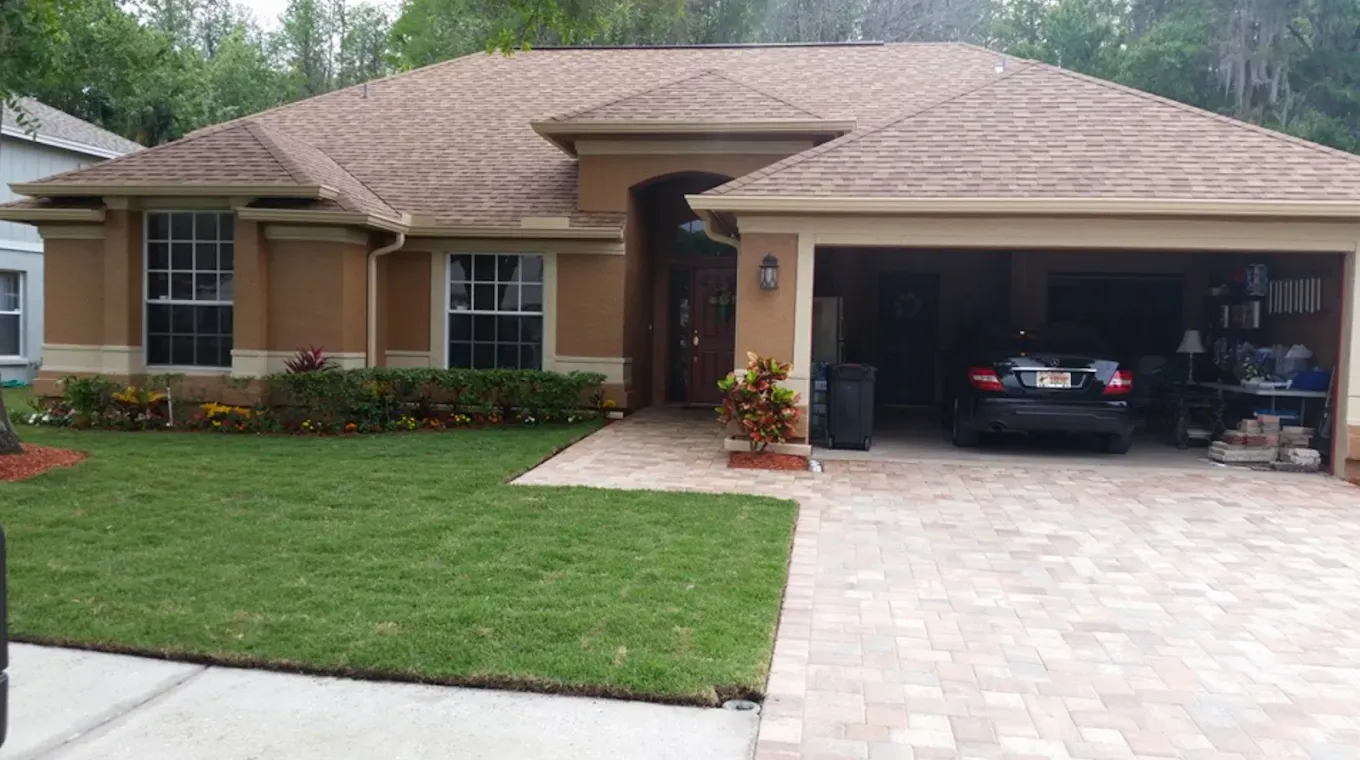 Tan house with a brick driveway and garage, green lawn, and flowering bushes.