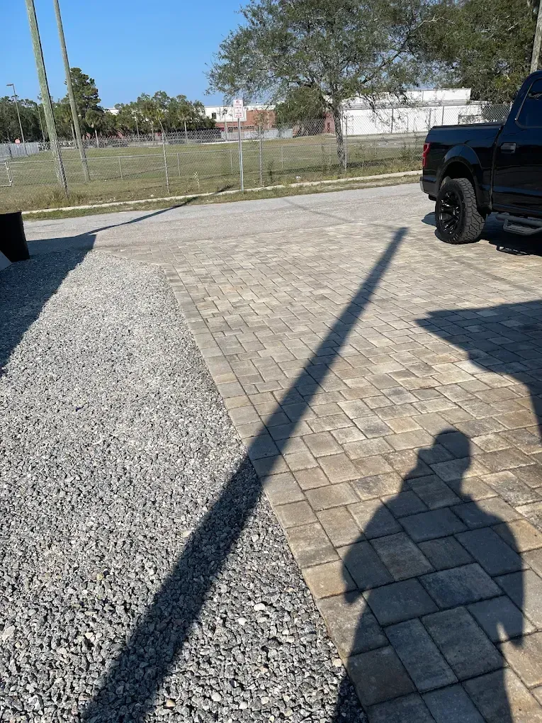Gravel pathway next to a brick paver driveway. A person's shadow stretches across the driveway from the right. A black truck is on the right side.