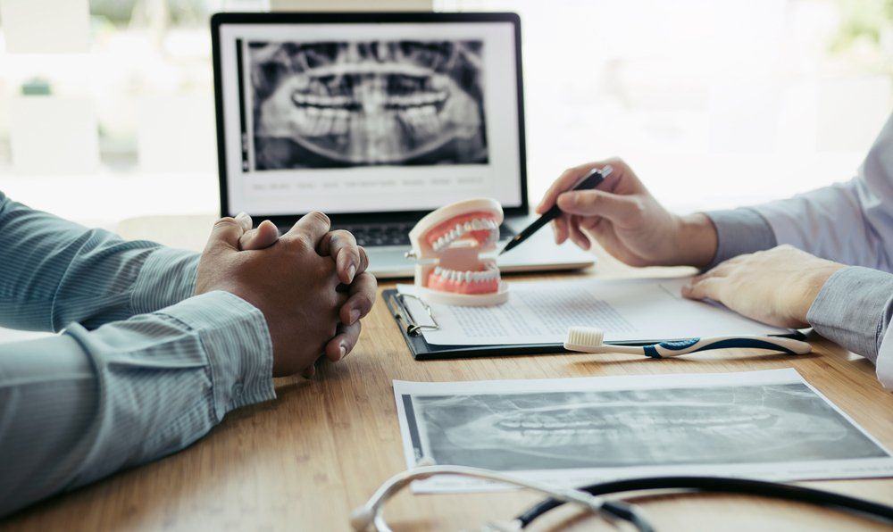 Dentist pointing to an x-ray on a laptop, explaining to a patient. Model teeth and other x-rays on desk.