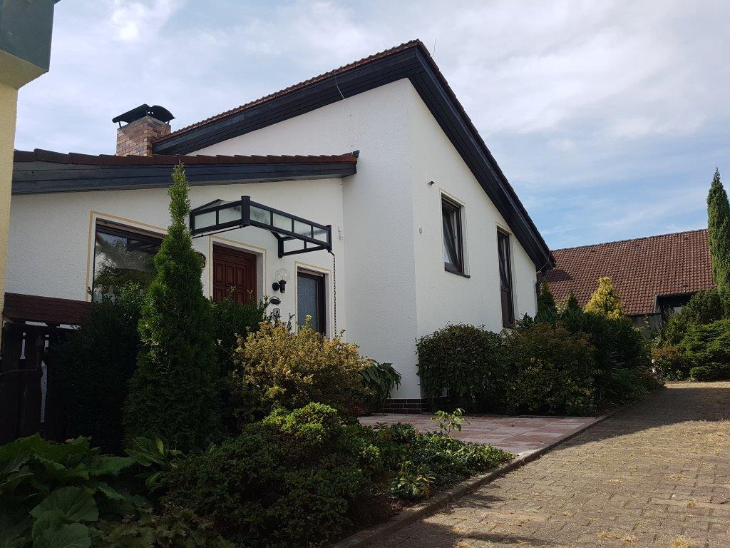 Ein weißes Haus mit braunem Dach ist von Bäumen und Büschen umgeben.