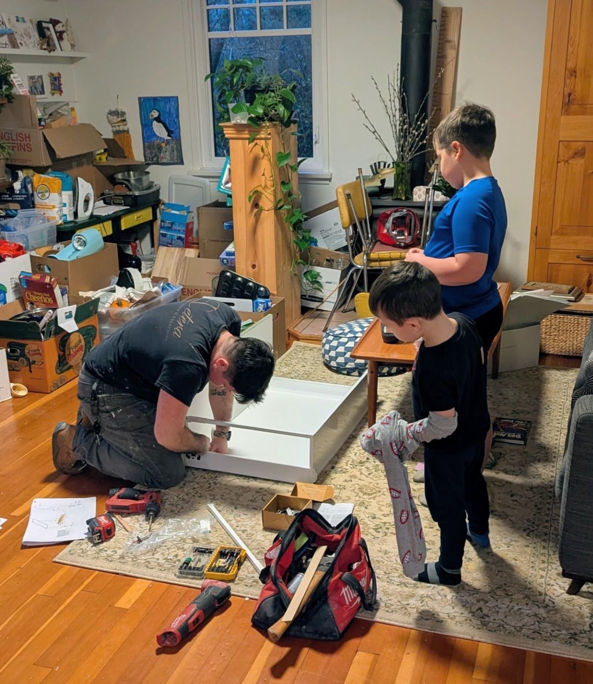 Children doing a craft project on the floor in a cluttered room, one kneeling and two standing nearby