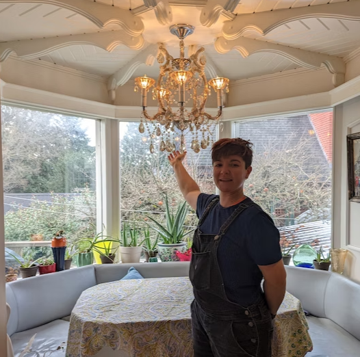 Person standing in a bright conservatory, pointing up at a gold chandelier above a round table.