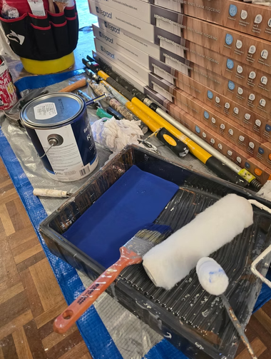 Paint supplies in a tray: blue paint, roller, brush, scraper, and caulk on a drop cloth next to stacked boards