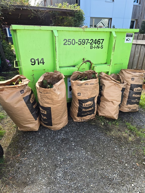 Five brown yard-waste bags in front of a bright green dumpster on a gravel driveway.
