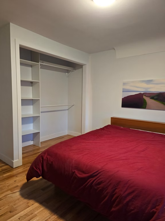Small bedroom with an open closet, hardwood floor, and a red bedspread beneath a wall painting.