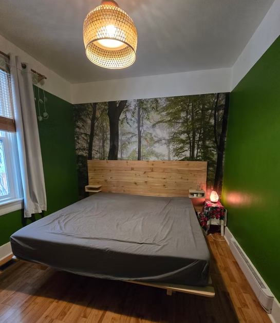 Bedroom with forest mural, wooden bed frame, gray bedding, green walls, and a woven ceiling light.