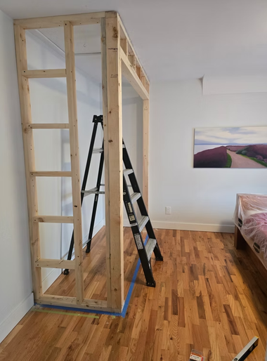 Wood-framed loft structure under construction in a bedroom, with a ladder leaning inside and a bed at right