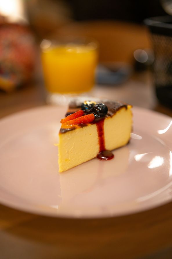 A slice of cheesecake on a white plate with a glass of orange juice in the background.