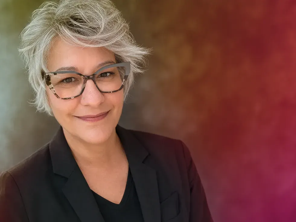 Smiling person with short gray hair and glasses in a black blazer against a warm, blurred background