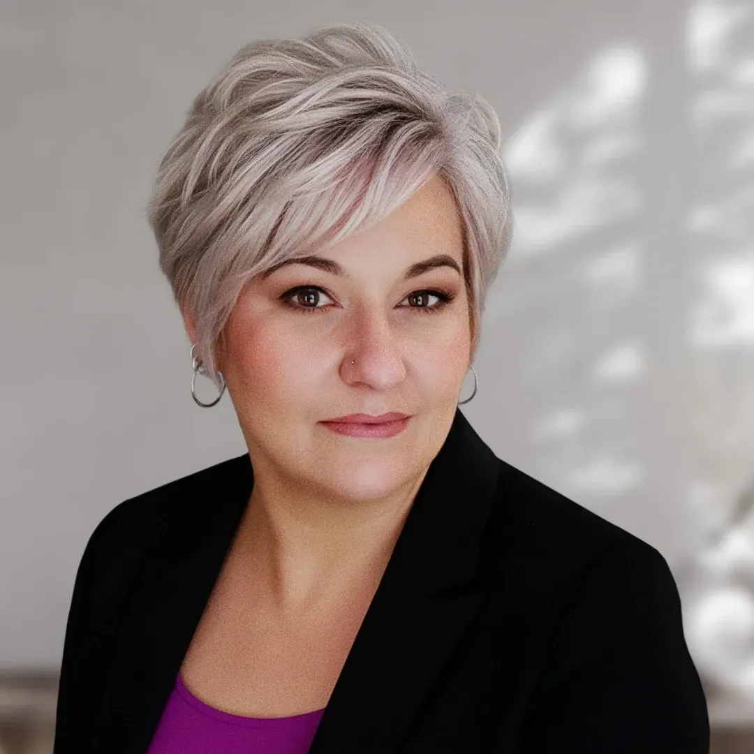 Portrait of a person with short silver hair in a black blazer and purple top, against a soft gray background