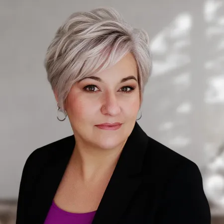 Portrait of a person with short silver hair in a black blazer and purple top, against a soft gray background