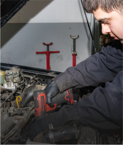 Mechanic working on a car engine, using a power tool while wearing black gloves. Red jack stands visible in the background | Allegiance Automotive
