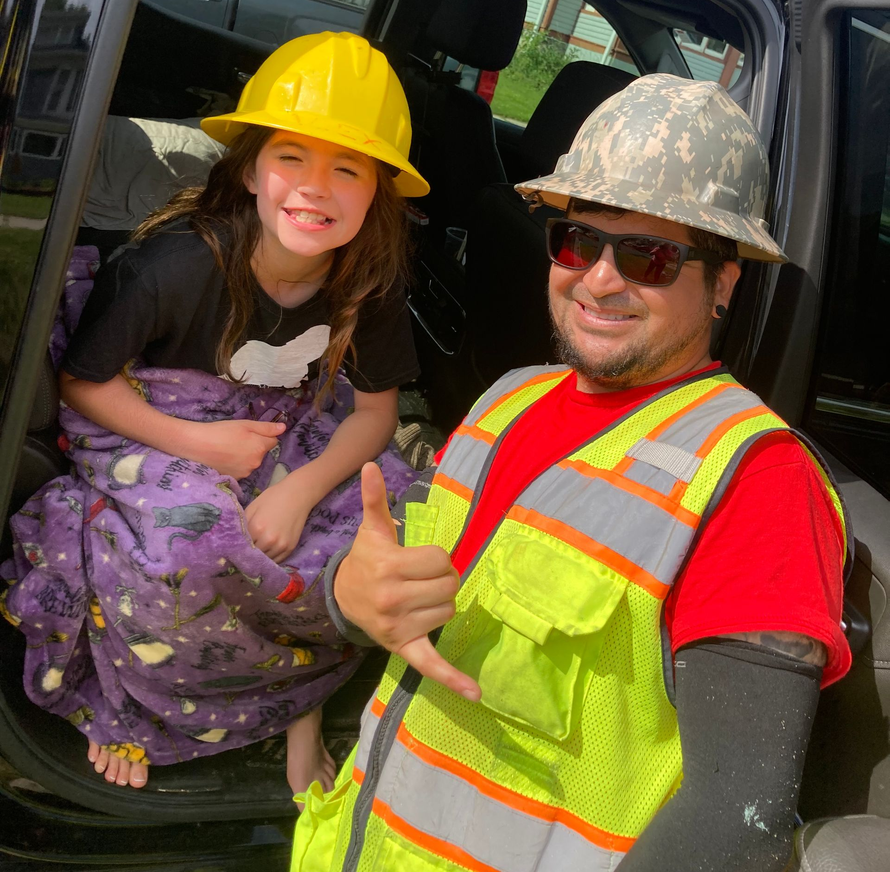 A man in a neon safety vest and hard hat makes a shaka sign next to a child in a hard hat wrapped in a purple blanket.
