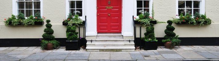 Three stone steps flanked by bushes in pots, leading to the red front door of a white house