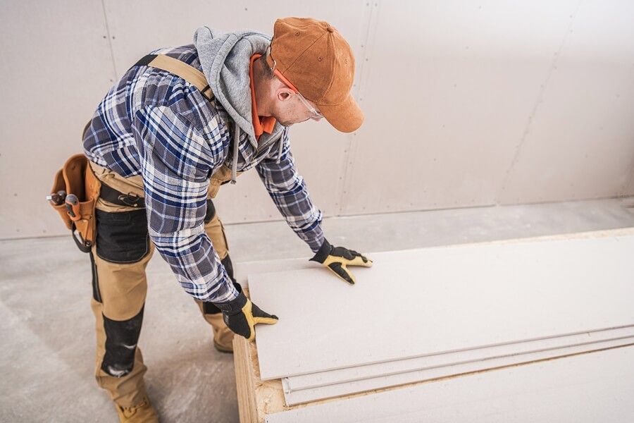 Construction worker handling drywall sheets in an unfinished room, wearing a cap, gloves, and overalls.