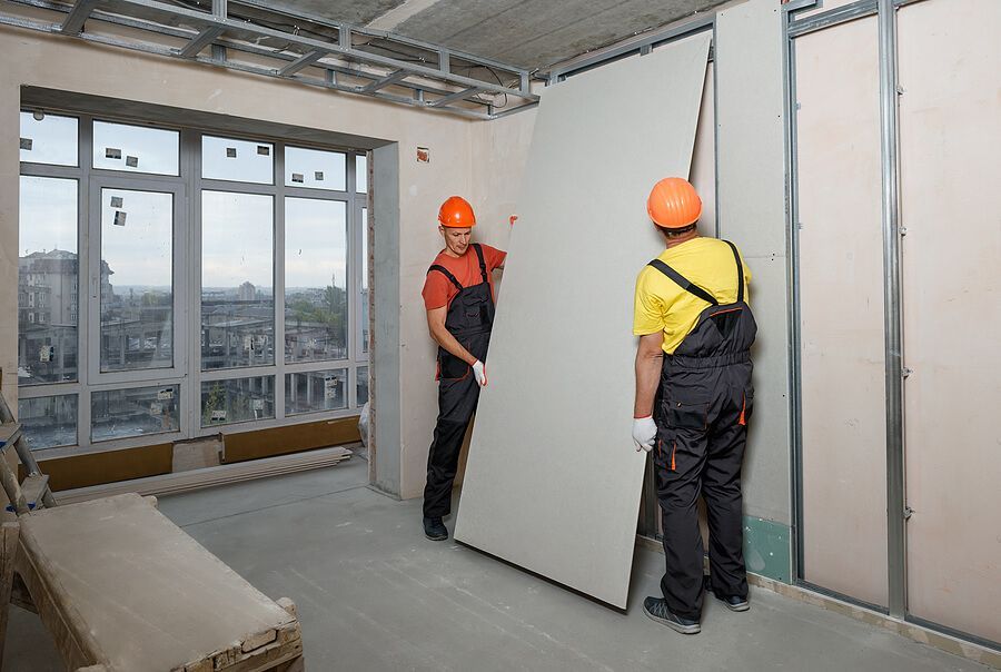 Two construction workers installing drywall in a building. They wear hard hats and overalls, working near a window.