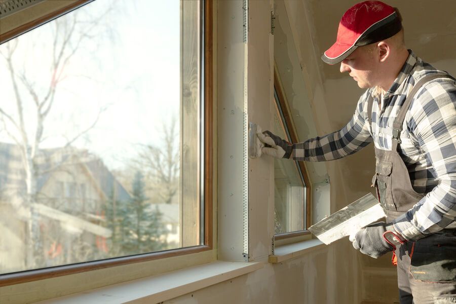 Man in cap and gloves applying plaster around a window frame in a room, using a trowel.