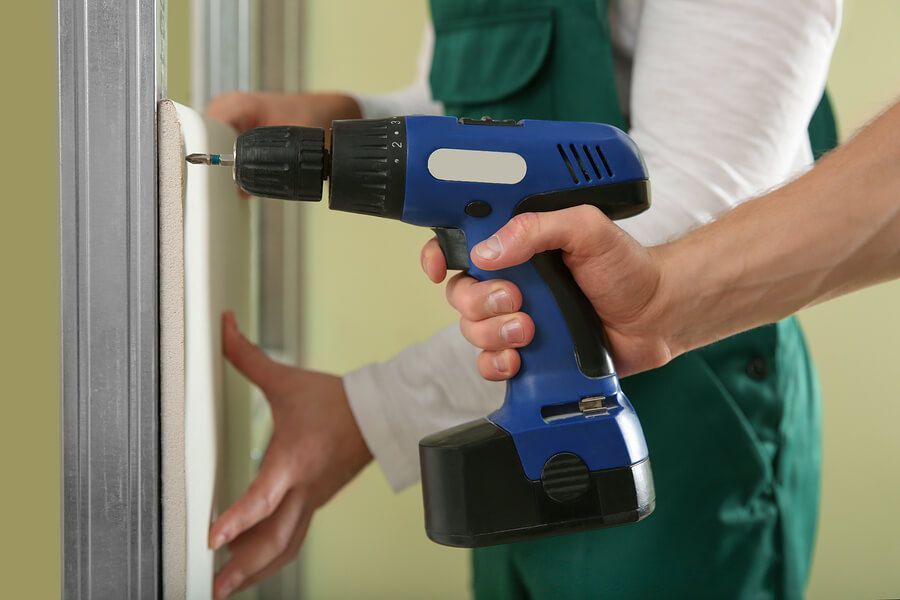 Person using a blue cordless drill to attach drywall to a metal stud. Another person's hand steadies the drywall.