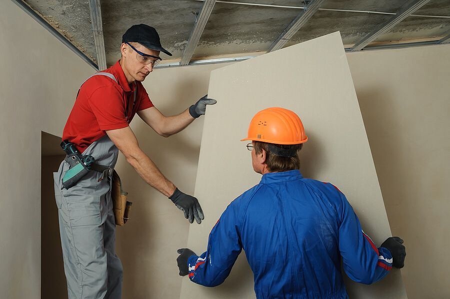 Two workers installing drywall in a room. One in red shirt and cap guides, the other in blue coveralls and helmet holds the sheet.