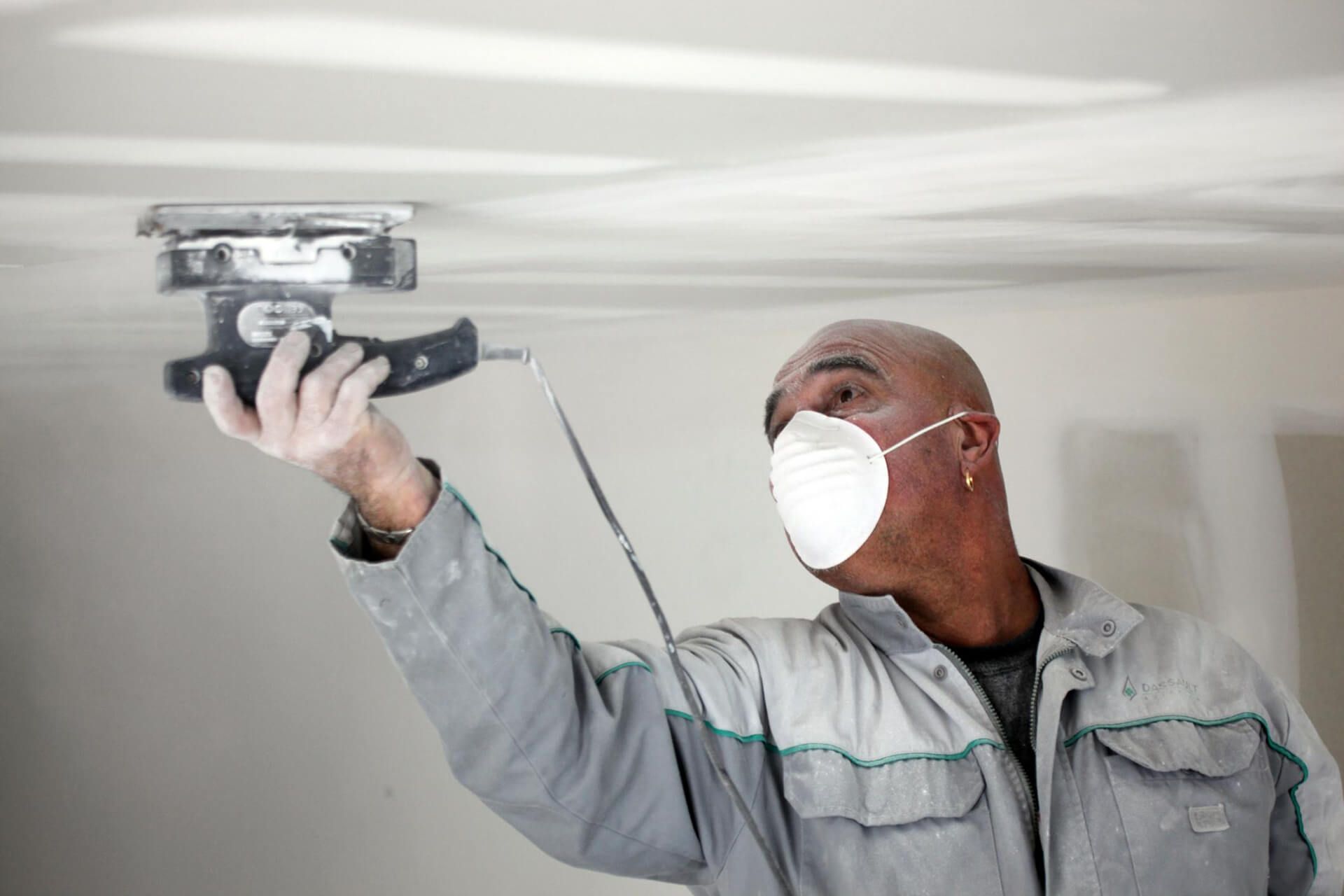 Person sands a ceiling with a power sander, wearing a dust mask, in a room.