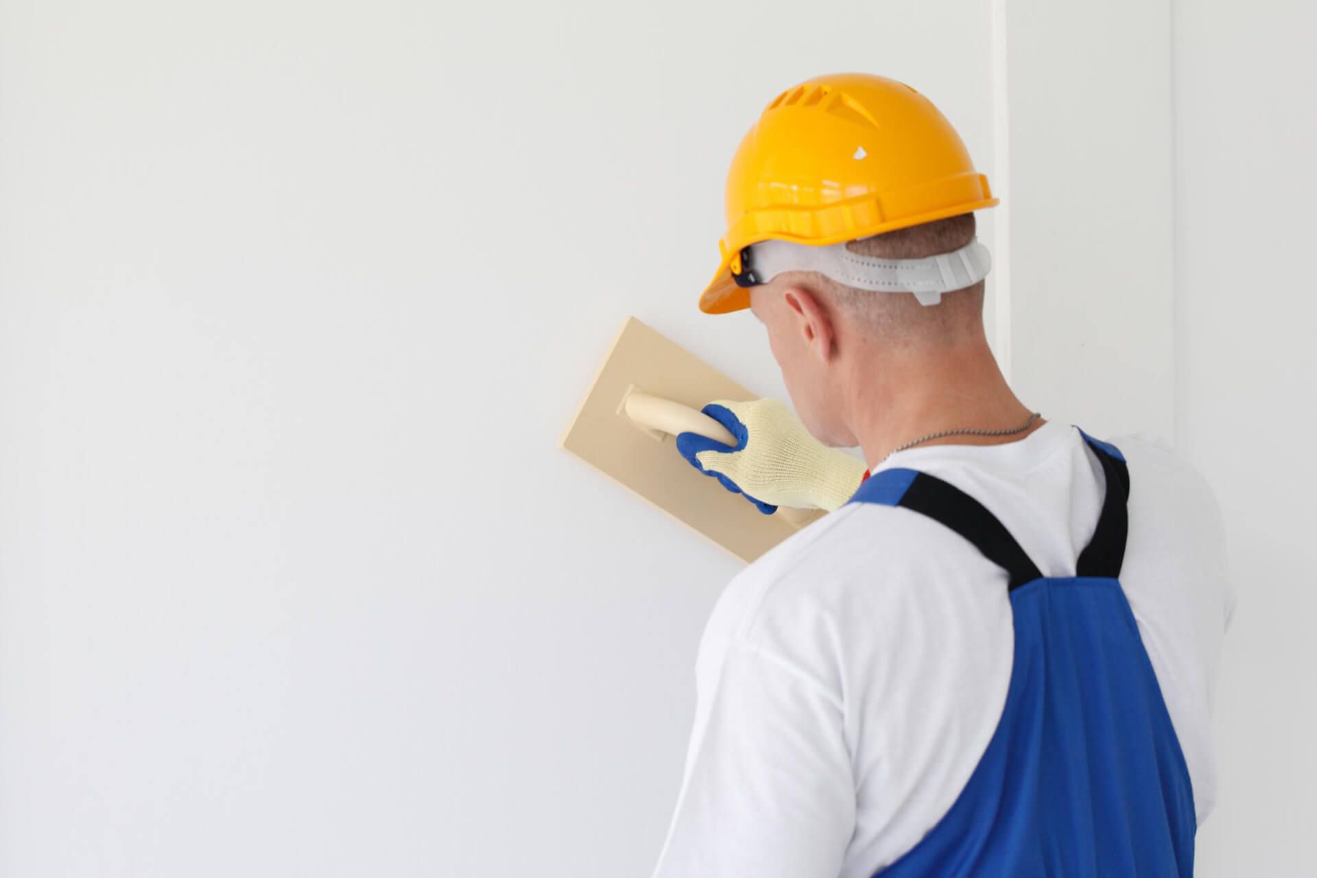 Person in yellow hard hat and blue overalls, using a trowel to smooth a white wall.