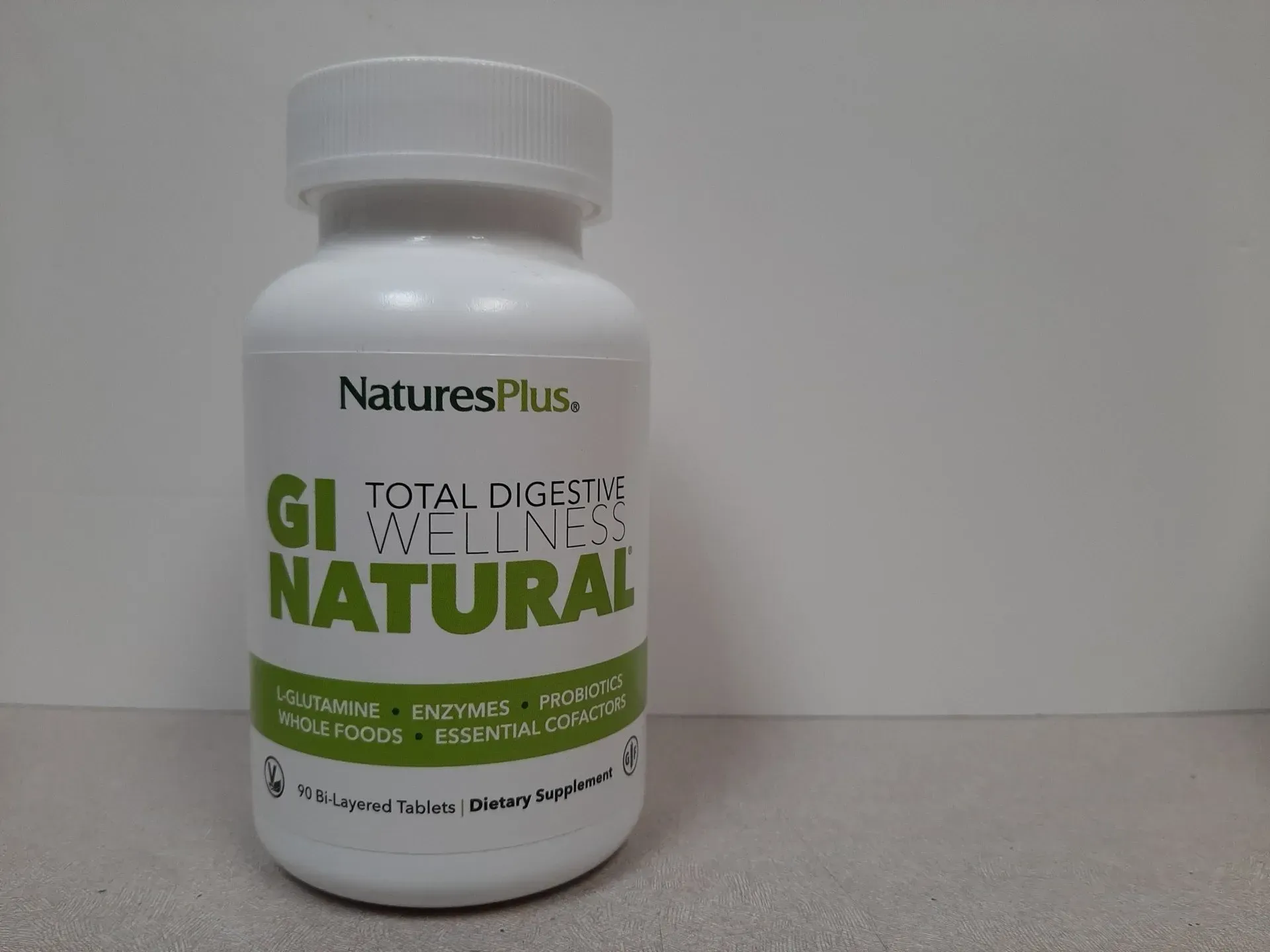 A white bottle of NaturesPlus GI Natural Total Digestive Wellness supplement standing on a surface against a white wall.