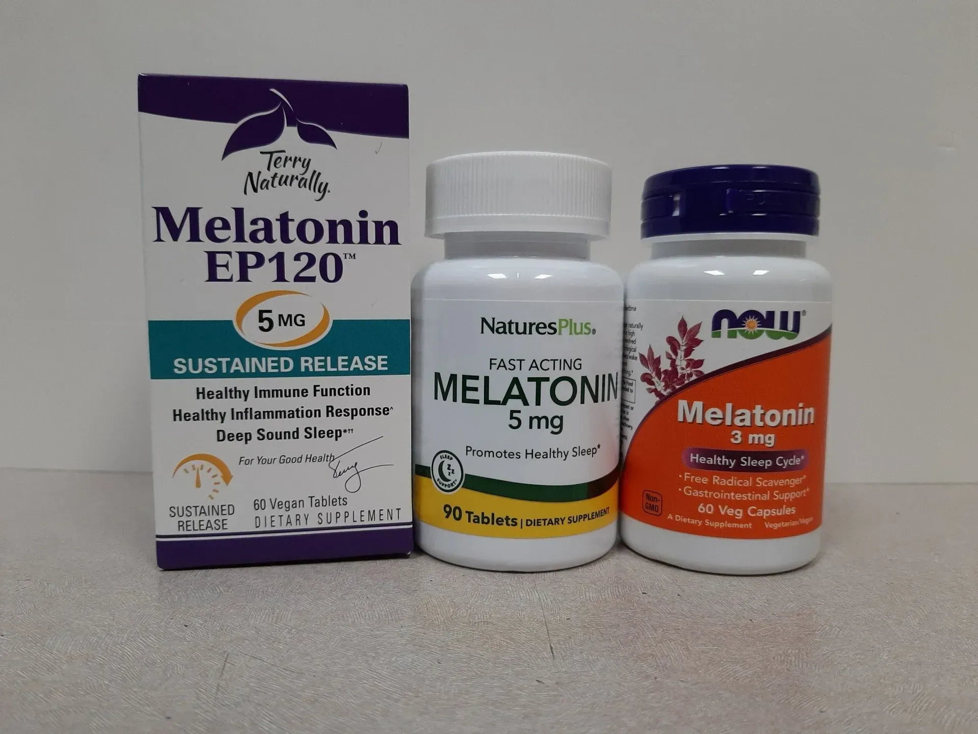 Three bottles of various melatonin supplements, including sustained-release and fast-acting, on a plain surface.