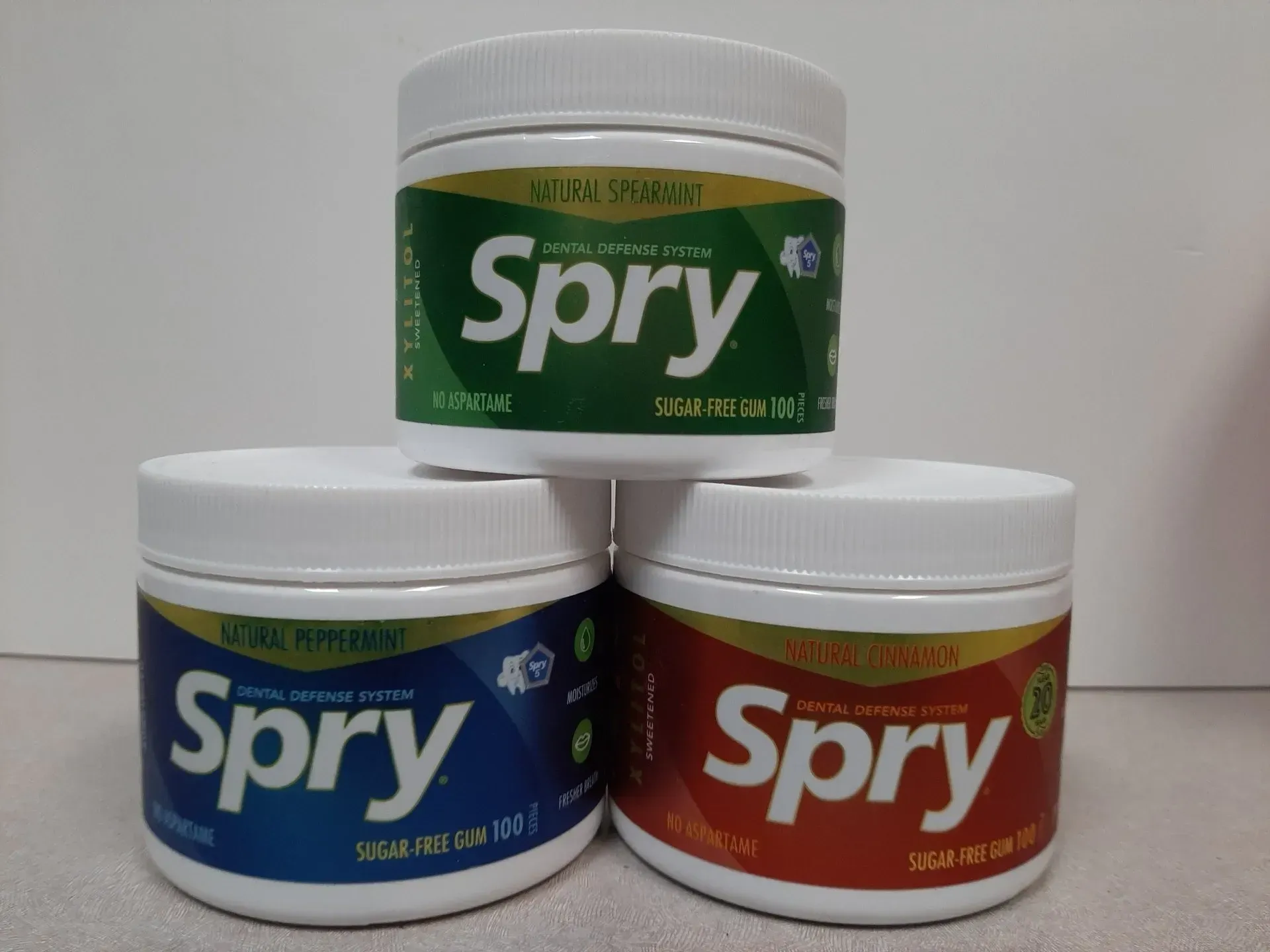 Three jars of Spry sugar-free gum stacked in a pyramid, featuring green, blue, and red labels on a white background.