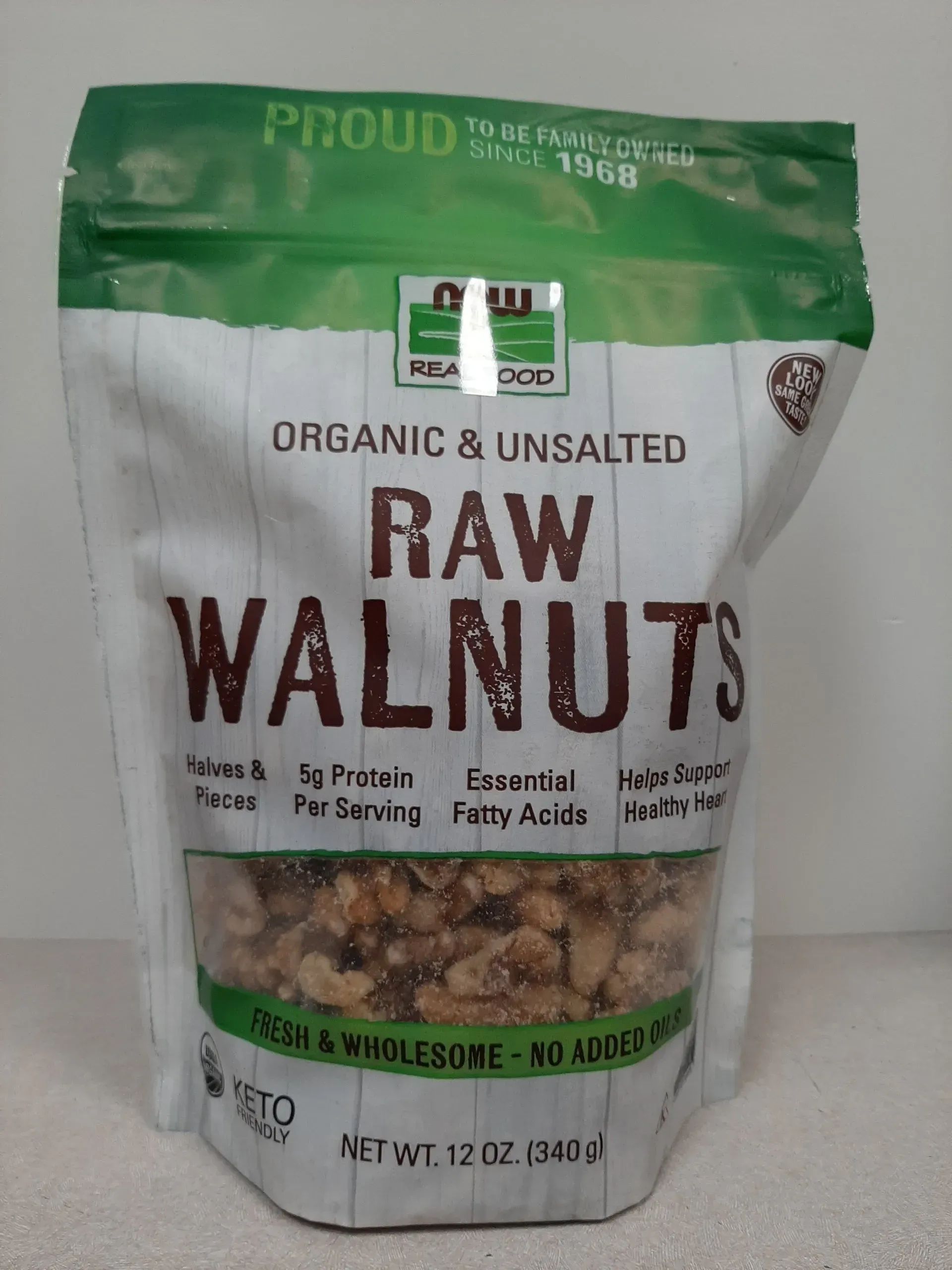 A 12oz bag of NOW Real Food organic, unsalted raw walnuts in white and green packaging against a plain background.