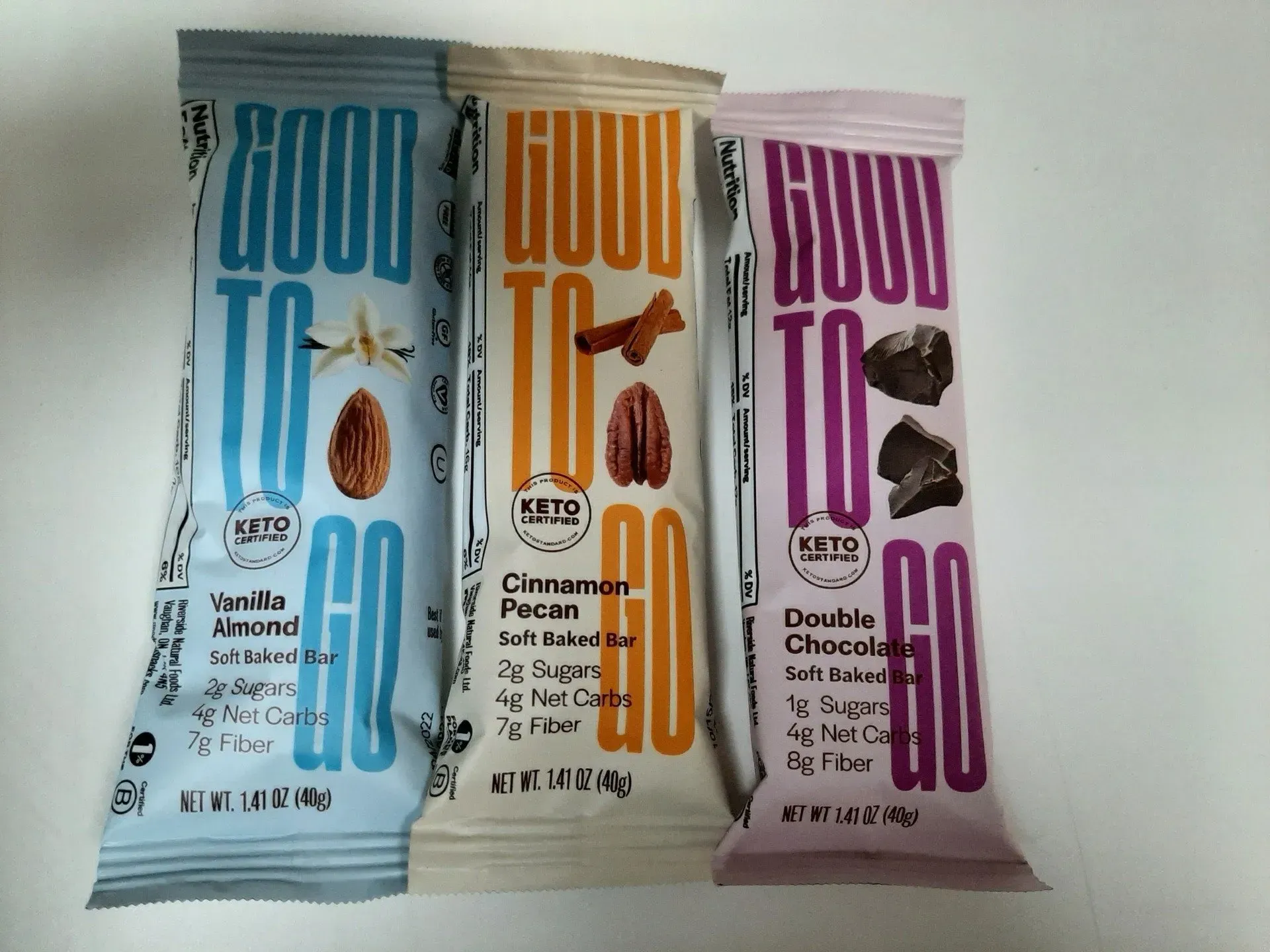 Three Good To Go keto snack bars in Vanilla Almond (blue), Cinnamon Pecan (tan), and Double Chocolate (purple) packaging.