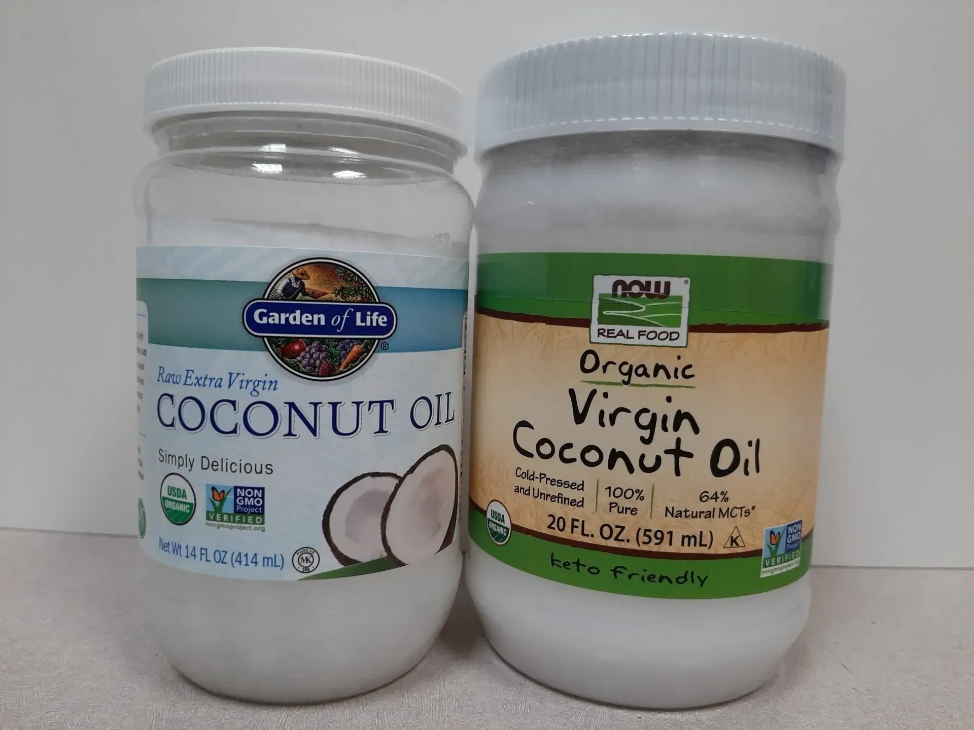 Two jars of organic virgin coconut oil from the brands Garden of Life and NOW Real Food sitting on a white surface.