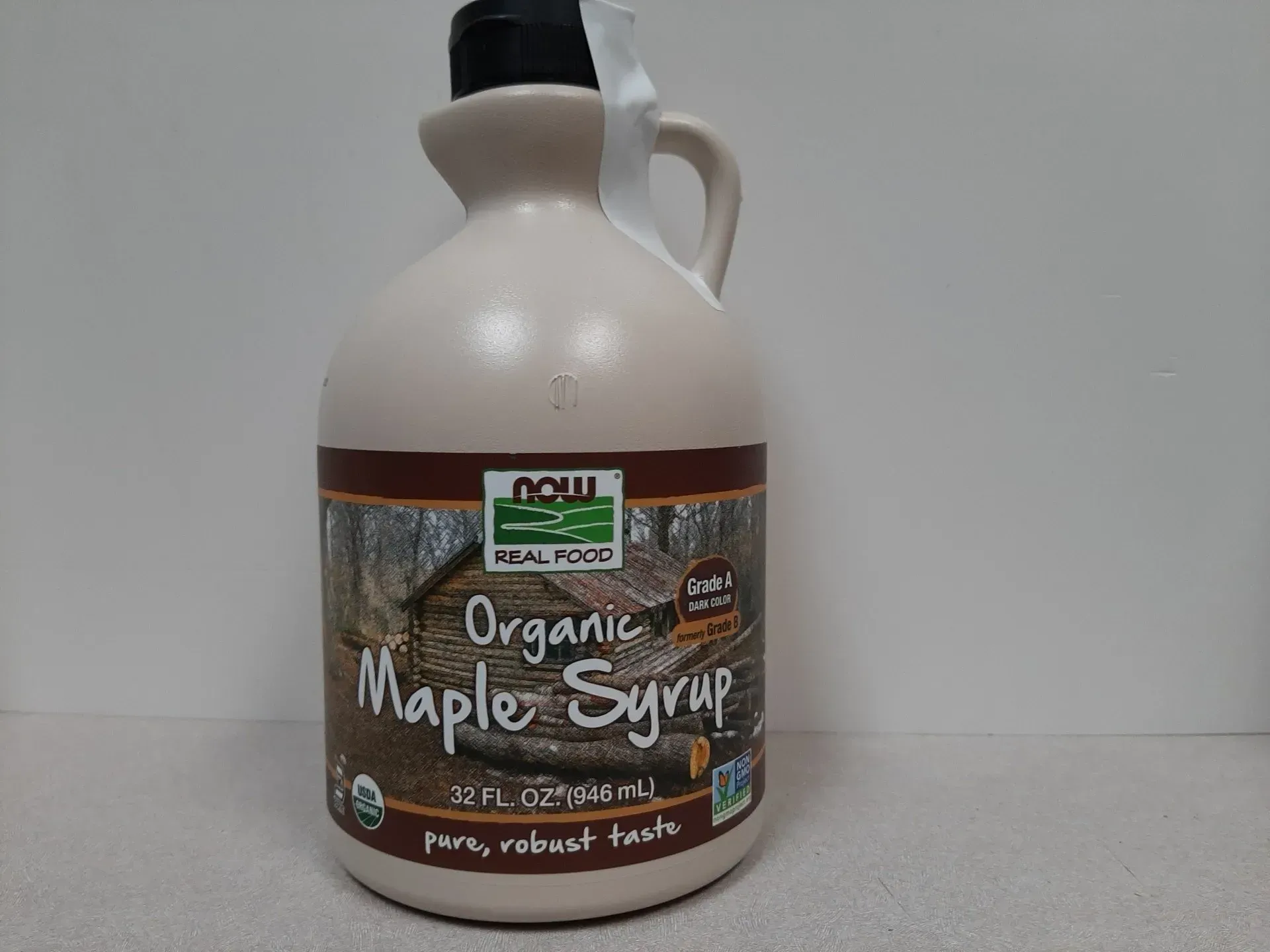 A large plastic jug of NOW Real Food Organic Maple Syrup with a tan label featuring a log cabin scene.