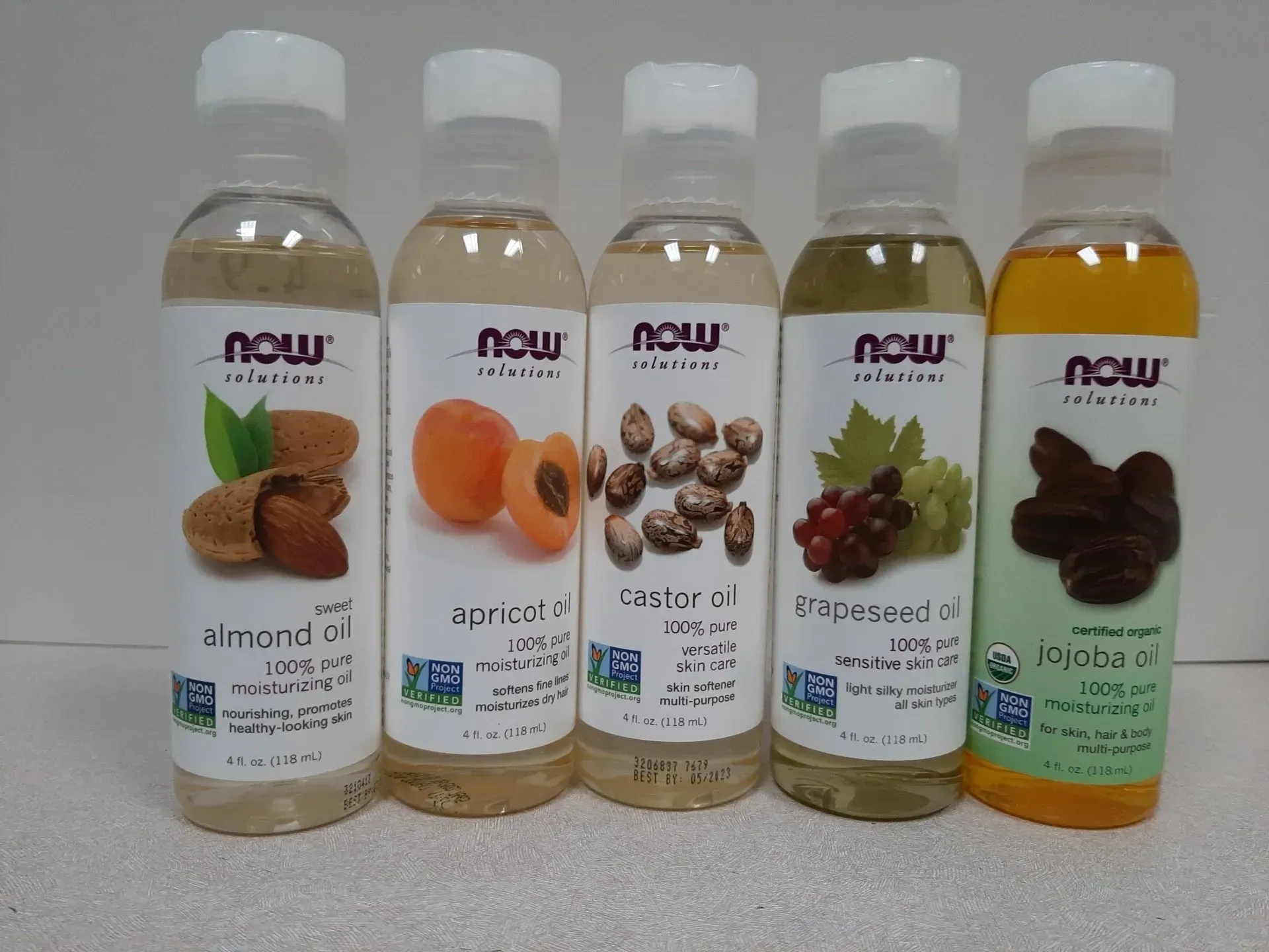 Five bottles of NOW Solutions oils, including almond, apricot, castor, grapeseed, and jojoba, lined up on a surface.