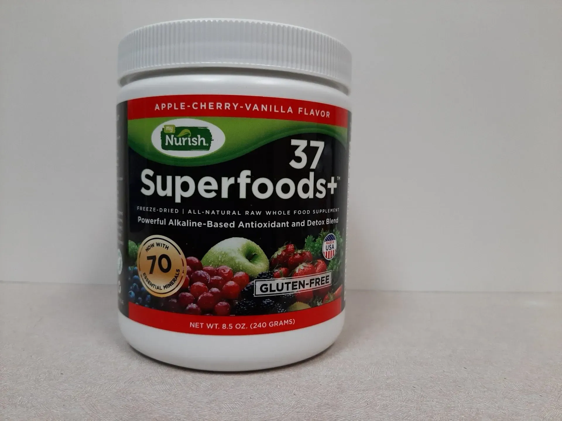 A white plastic tub of Nurish 37 Superfoods+ powder, apple-cherry-vanilla flavor, centered on a light surface.