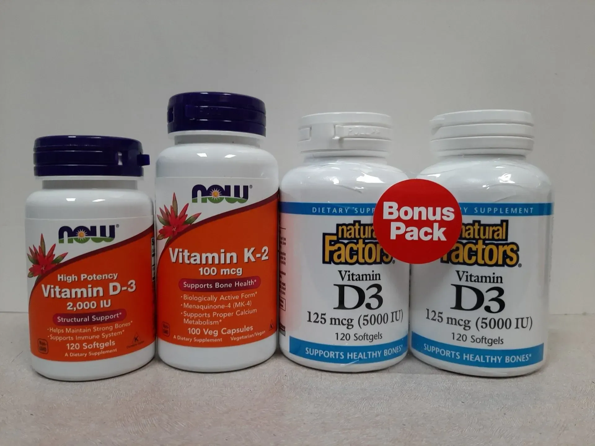 Four bottles of Vitamin D3 and K2 dietary supplements from NOW and Natural Factors brands lined up on a white surface.