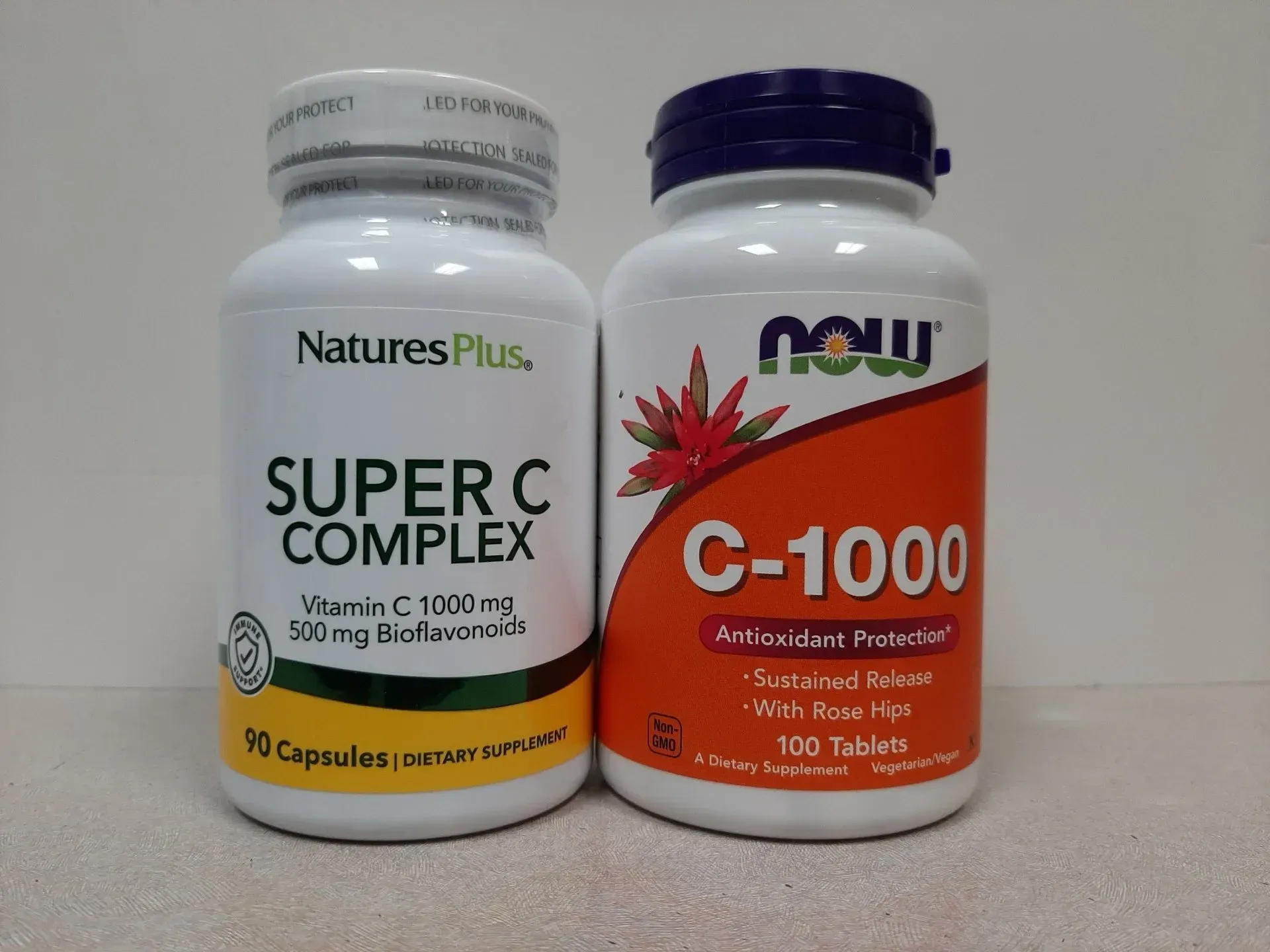 Two bottles of Vitamin C supplements, NaturesPlus Super C Complex and NOW C-1000, sit side-by-side on a light surface.