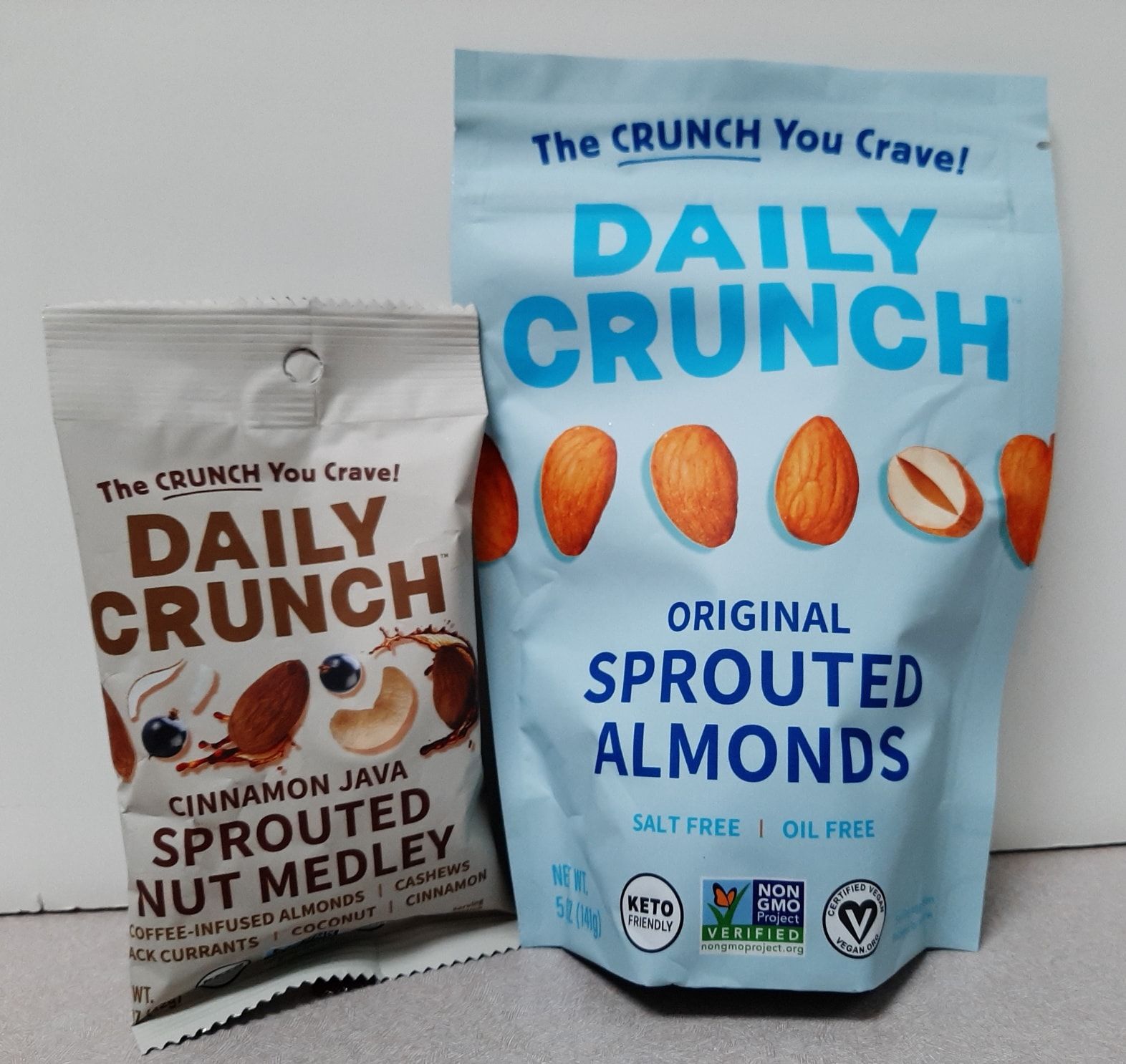 A light blue bag of Daily Crunch Original Sprouted Almonds next to a smaller cream-colored bag of Cinnamon Java Nut Medley.