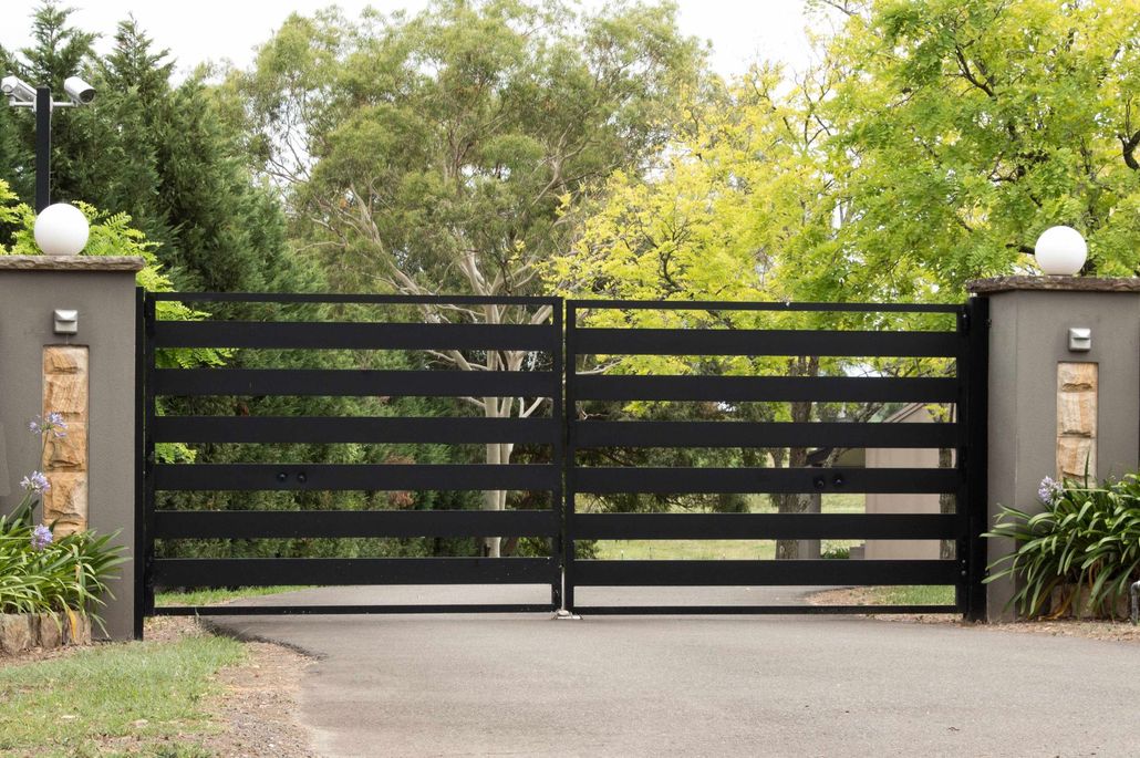 Black metal driveway gate between stone pillars and landscaped greenery, with road in front