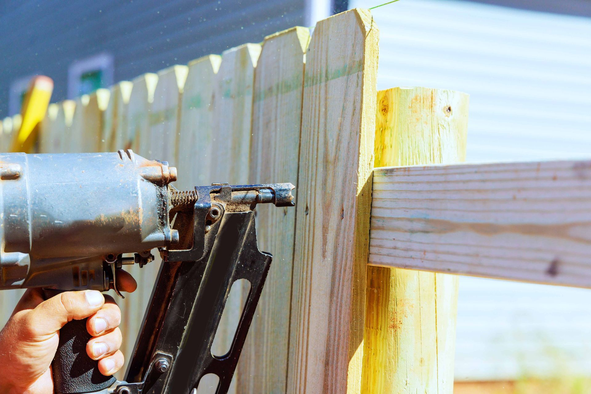 Wooden privacy fence with tan posts lining a roadside, with trees behind it. Hand holding a nail gun fastening a wooden fence board outdoors