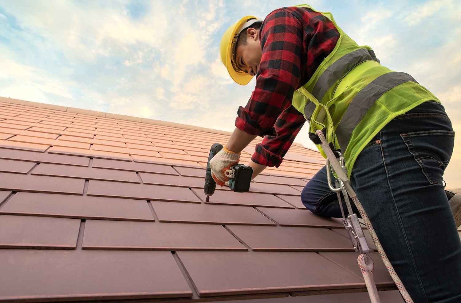 A residential roofing contractor installing shingles with a power drill on a house roof. A residential roofing contractor installing shingles with a power drill on a house roof.