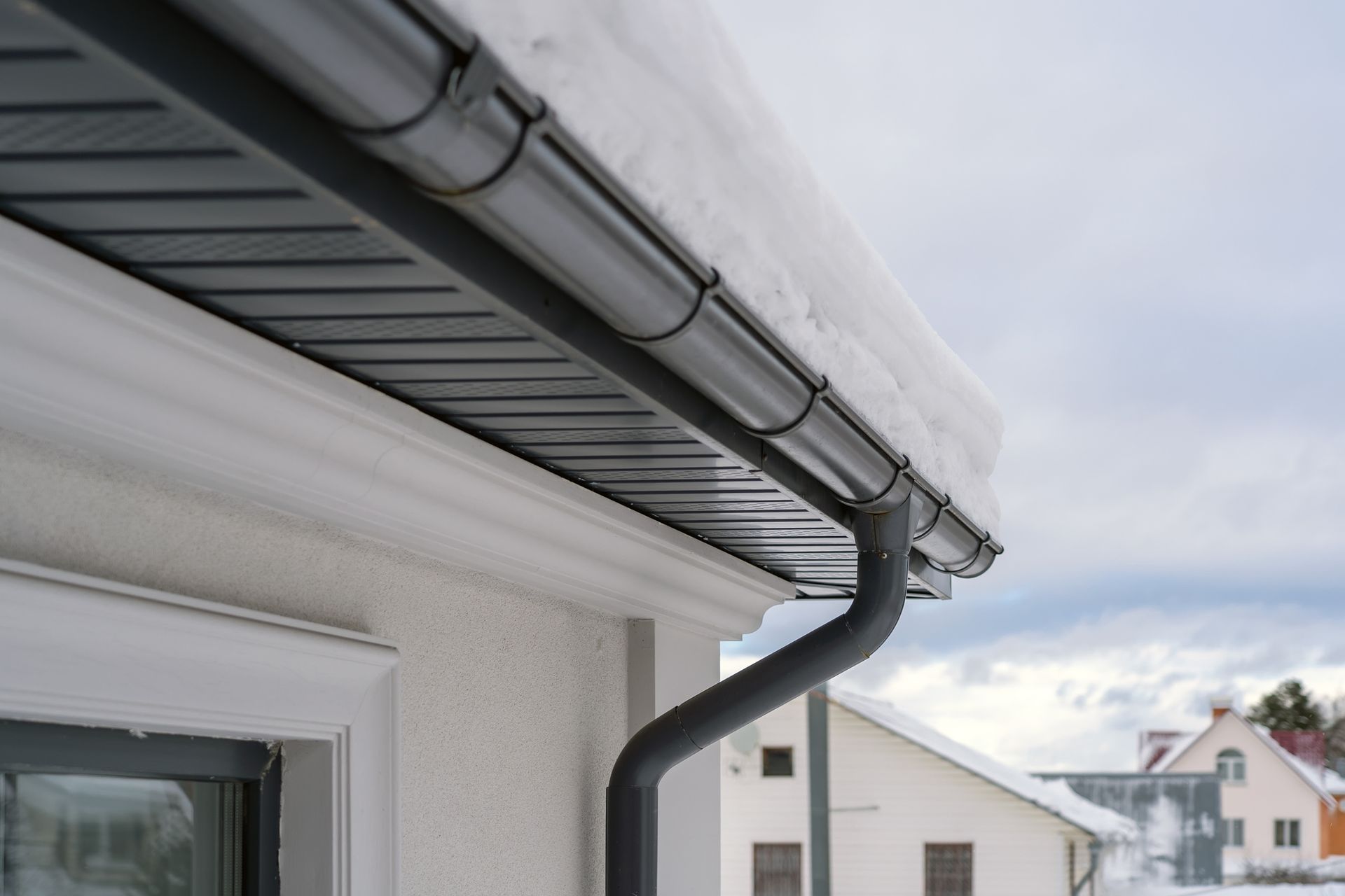 Snow-covered roof with black gutters and downspout on a white house, cloudy sky in the background. Snow-covered roof with black gutters and downspout on a white house, cloudy sky in the background.