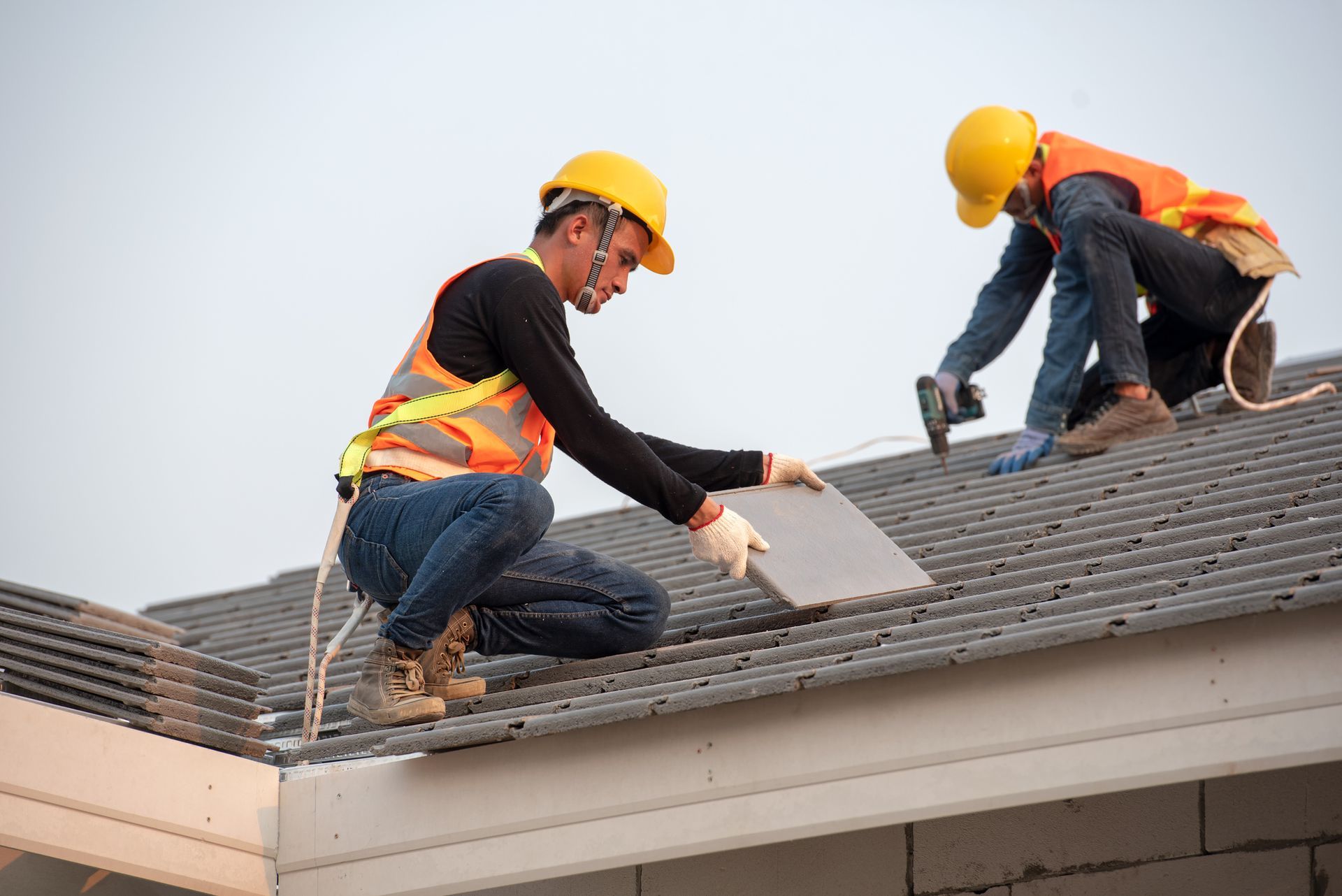 Two roofers in hard hats and safety vests installing tiles on a gray roof.