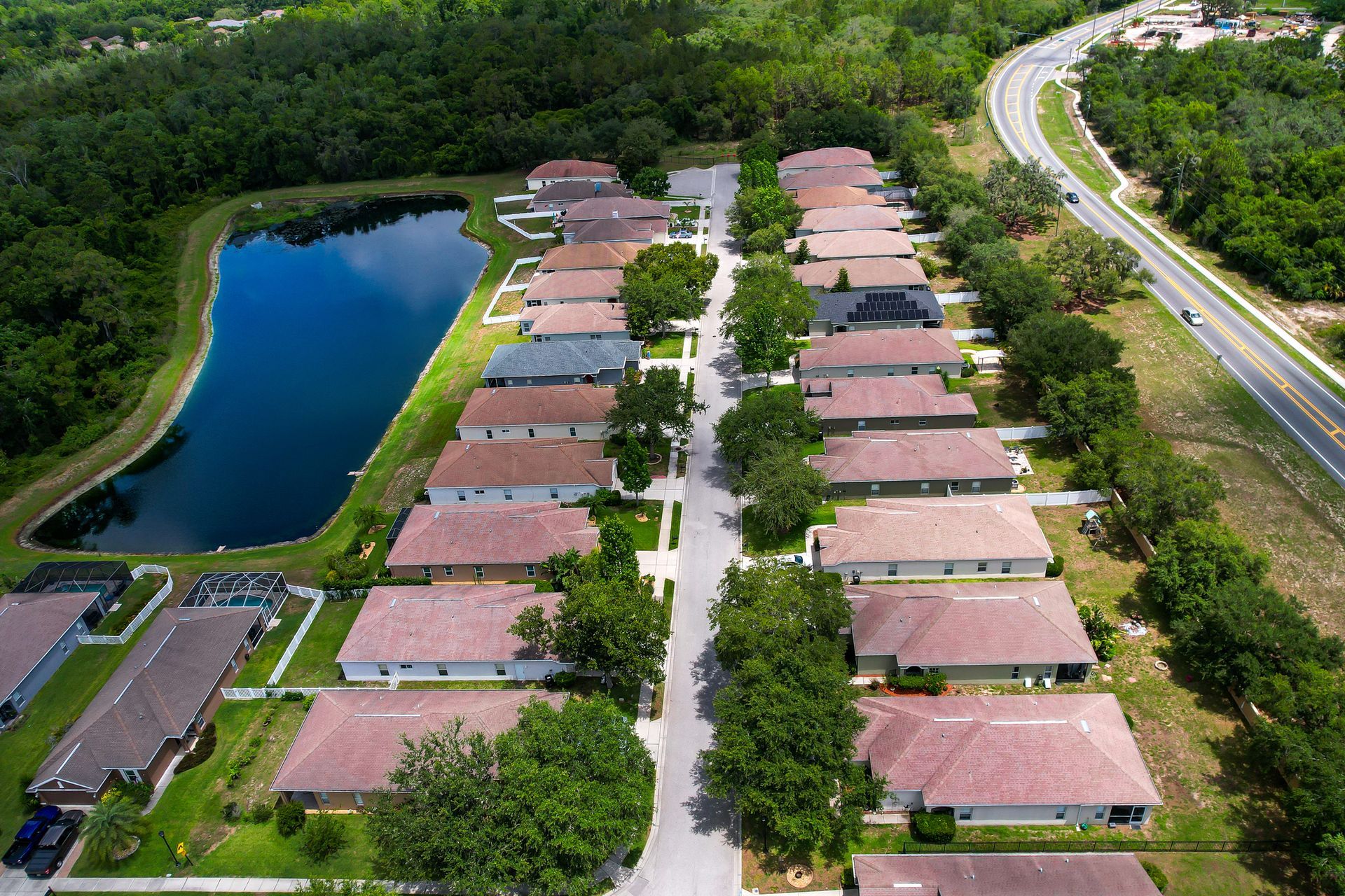 Aerial view of a suburban neighborhood with houses, a lake, road, and trees.